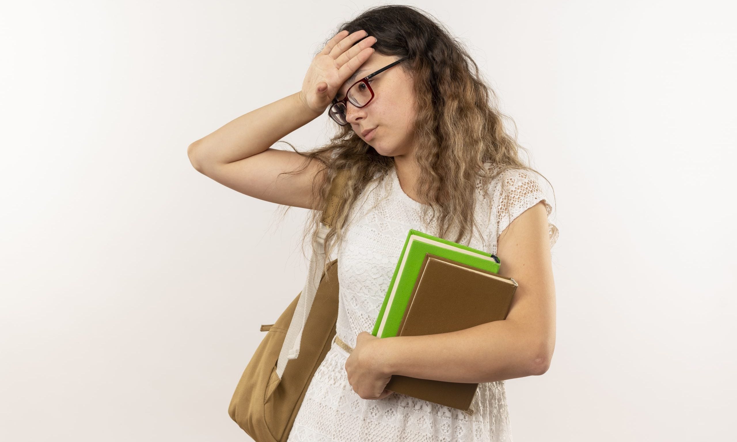 tired young pretty schoolgirl wearing glasses and back bag holding books looking down putting hand on head isolated on white background with copy space